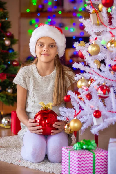 Una chica con una bola roja se sienta junto al árbol de Navidad, en el fondo hay linternas borrosas — Foto de Stock