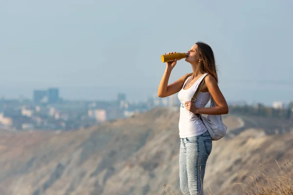 Girl drinks from a bottle of water, standing against the backdrop of a mountain and city landscape — Stock Photo, Image