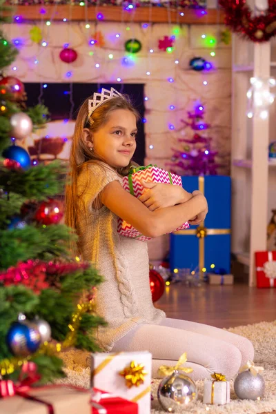 Niña de diez años se sienta en el árbol de Navidad y abraza un regalo —  Fotos de Stock