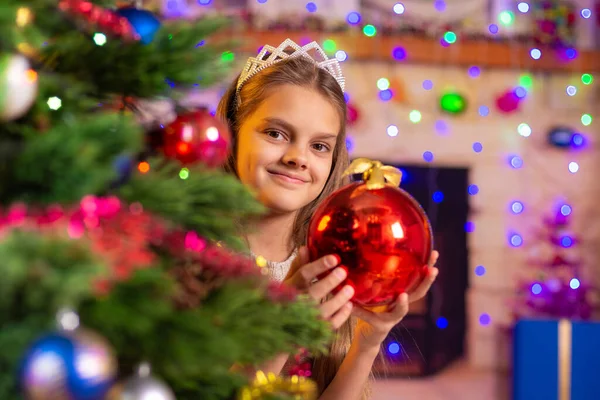 Chica feliz se asoma por detrás de un hermoso árbol de Navidad con una pelota en sus manos —  Fotos de Stock