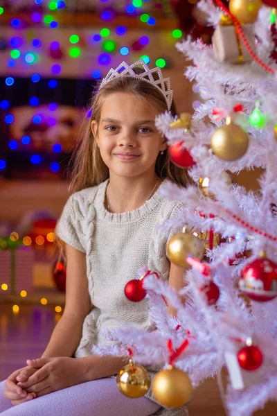 Hermosa chica se sienta en el árbol de Navidad sobre el fondo de luces borrosas — Foto de Stock