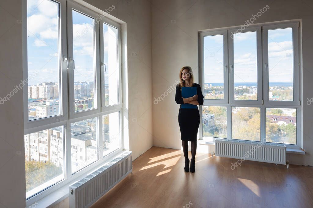 The girl with the folder stands in the middle of a large spacious room with large windows