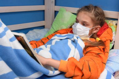 Tıbbi maskeli hasta kız yatakta uzanıyor ve kitap okuyor.