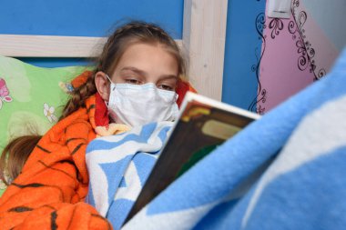 Genç kız tıp maskesiyle yatakta uzanıyor ve kitap okuyor.