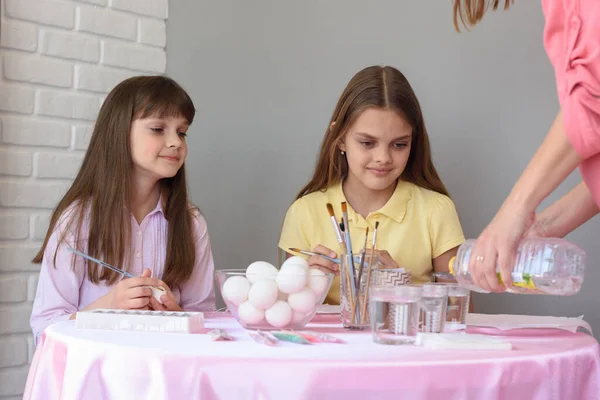 Mom pours vinegar into glasses with food coloring, children at the table prepare to paint eggs