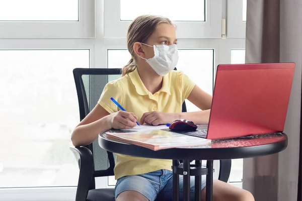 A girl in a medical mask on quarantine in isolation isolates herself remotely from her lessons and distracted, looked out the window