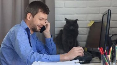 Genç bir adam evde çalışıyor ve telefonda konuşuyor, arka planda komik bir kedi.