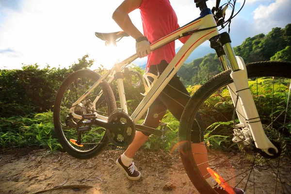 Young woman cyclist carrying mountain bike climbing on summer forest trail