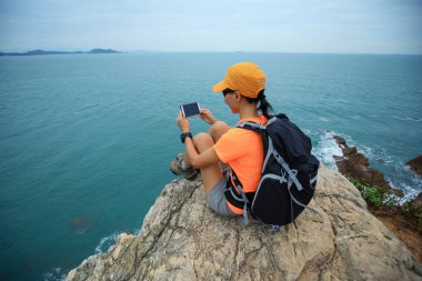 Genç kadın uzun yürüyüşe çıkan kimse alarak fotoğraf ile deniz cliff üzerinde otururken smartphone