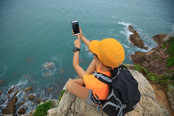 坐在海边悬崖上的年轻女子徒步旅行者使用智能手机 — 图库照片