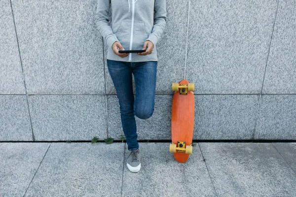 都市の壁にもたれながらスマート フォンを使用して女性のスケートボーダー — ストック写真