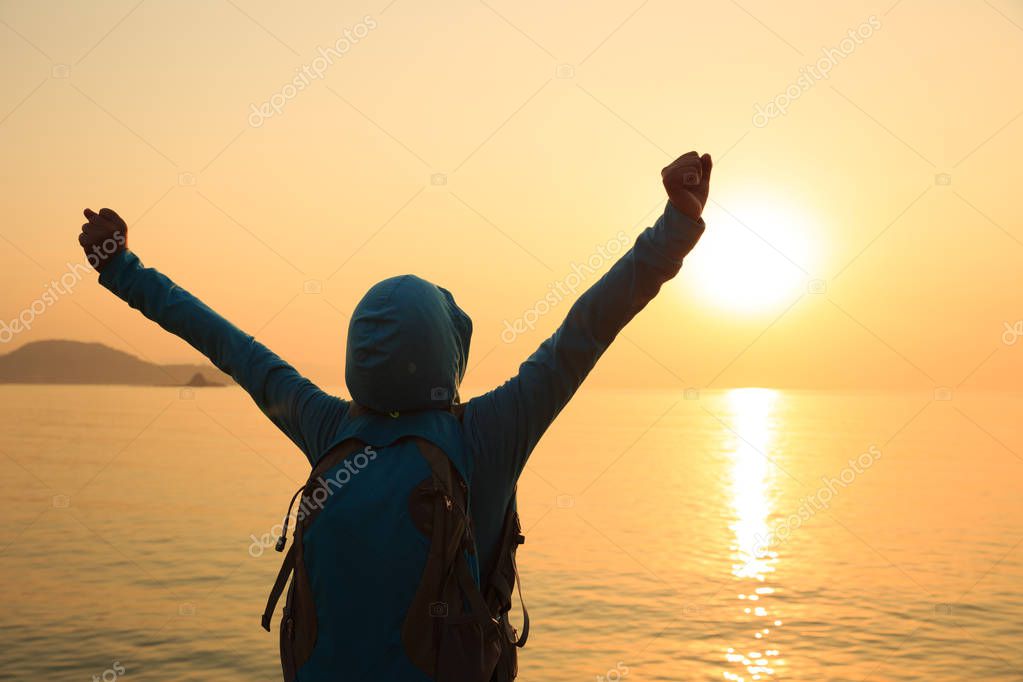 Silhouette of cheering female hiker with backpack standing on sunrise windy coast