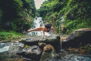 Genç kadın ormanda şelalenin yanında yoga yapıyor.