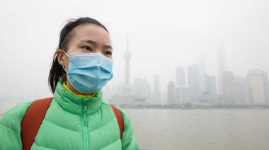 Endişeli yeşil ceketli ve maskeli Çinli kadın kirlilik sisi ile şehre bakıyor..