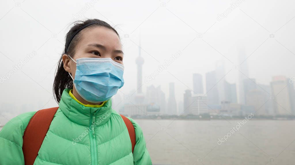 Worried Chinese woman in green jacket and face mask looking away at city with pollution fog.
