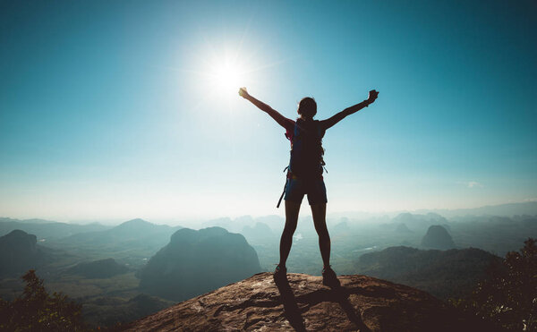 Cheering female hiker with backpack enjoying view on sunrise at mountain top cliff edge