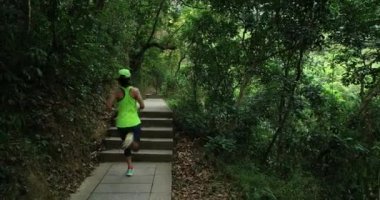 Spor giyimli genç bir kadın tropik orman yolunda koşuyor.