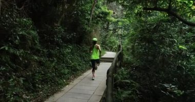 Spor giyimli genç bir kadın tropik orman yolunda koşuyor.