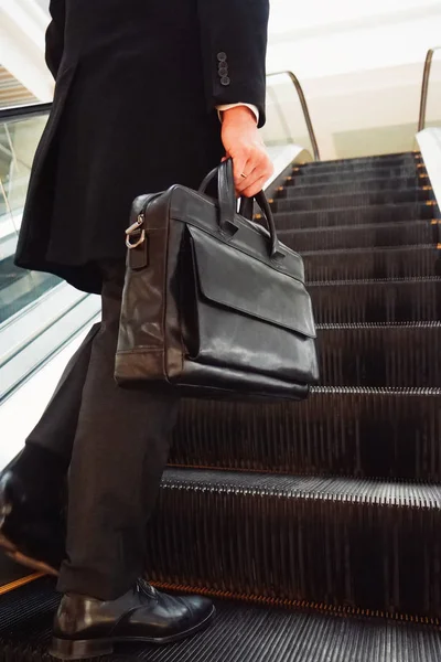 Un hombre en un traje de negocios y abrigo largo con un maletín de cuero negro sube a la escalera mecánica — Foto de Stock