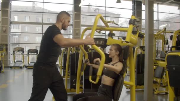 Mujer joven con instructor usando máquina de prensa de pecho en el gimnasio — Vídeos de Stock