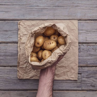 Bir erkek el, bir torba patatesi taşralı bir arka planda tutuyor.