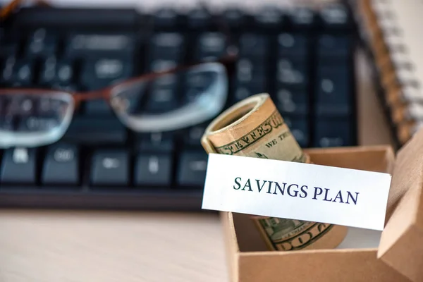 Words savings plan print on pieces of paper, money banknotes dollars in craft box, glasses and on table.