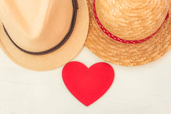 Two straw panamas hat man and woman close up and red wooden heart on table. The concept of a couple in love, a romantic holiday for two.