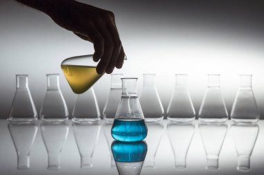 El sarısını mavi sıvıya döküyor bilimsel laboratuvar bardağı erlenmeyer şişesinde yansıtıcı yüzeyde cam ekipmanla birlikte..
