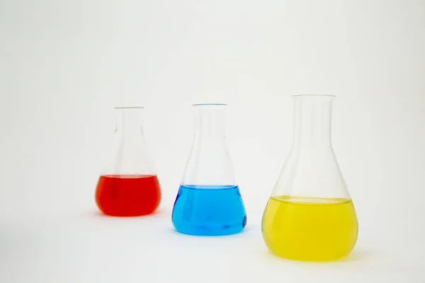 Beyaz zemin üzerinde mavi, sarı ve kırmızı sıvıyla dolu bilimsel laboratuar camı Erlenmeyer şişesi.. — Stok fotoğraf