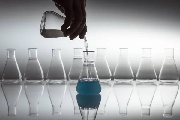 Verser à la main du blanc dans un liquide bleu dans une fiole d'erlenmeyer en verre de laboratoire scientifique avec un équipement de verrerie sur une surface réfléchissante. — Photo