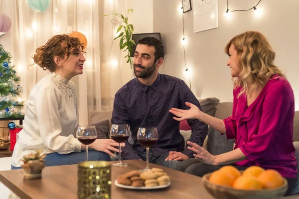 Friends in living room having fun by the Christmas tree, speaking an smiling with glass of wine.