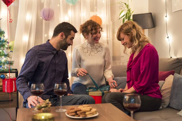 Friends enjoying moment of sharing and opening presents by Christmas tree in living room.