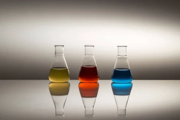 Yansıtıcı yüzeyde sarı, kırmızı ve mavi sıvıyla dolu bilimsel laboratuar camı Erlenmeyer şişesi.. — Stok fotoğraf