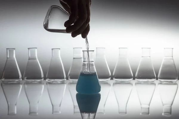 Bilimsel laboratuvar bardakları Erlenmeyer şişesinde yansıtıcı yüzeyde cam ekipmanla mavi sıvıya beyazı döküyorum.. — Stok fotoğraf