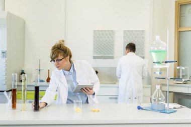 Gözlük takan kadın kimya bilimci laboratuvarda kenevir CBD ve CBDA yağlarıyla çalışıyor. Lateks eldiven, cam tüpler ve erlenmeyer şişeleri kullanıyor. Esrar tohumlarından alınan sağlık hizmetleri eczanesi.