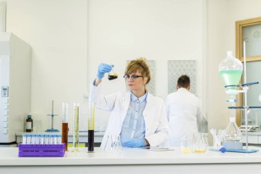 Gözlük takan kadın kimya bilimci laboratuvarda kenevir CBD ve CBDA yağlarıyla çalışıyor. Lateks eldiven, cam tüpler ve erlenmeyer şişeleri kullanıyor. Esrar tohumlarından alınan sağlık hizmetleri eczanesi.