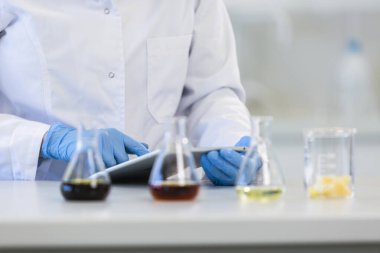 Tıbbi eldivenli eller laboratuvar masasının üzerinde beyaz tablete yazı yazıyor. Kenevir tohumu, marihuana tomurcuğu, CBD ve CBDA yağları masada. Esrar eczacılık sağlık hizmeti kavramı.