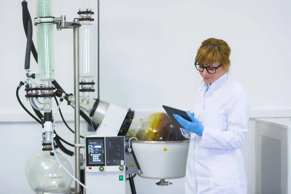CBDA yağ çıkarma işlemi sırasında laboratuvarda siyah tablet üzerinde yazan bir bilim adamı. Lastik eldiven takıyor ve rotavapor denilen yeşil yoğunlaştırıcılı döner buharlaştırıcının yanında duruyor..