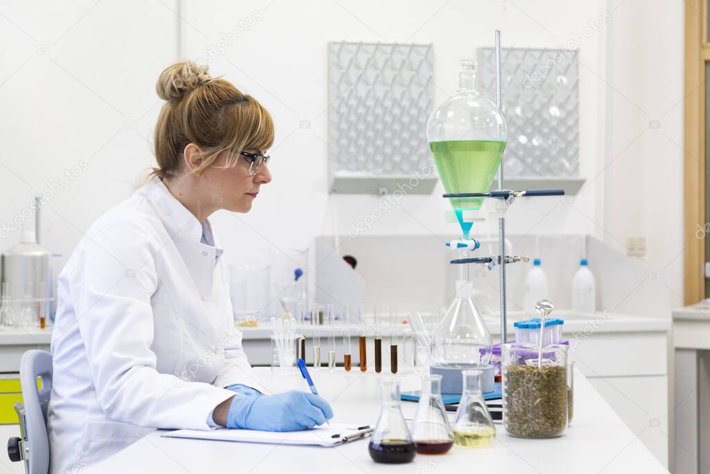 Female scientist wearing glasses and latex gloves writes measurement results with blue pen. Turquoise liquid in pear shaped separatory funnel is in background. Hemp seeds and cbd oils are on table.