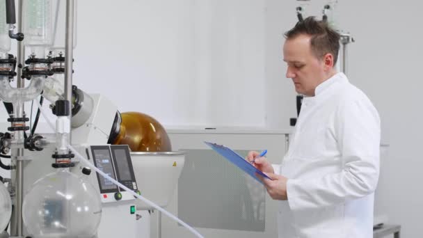 Cbd油抽出中に研究室で回転蒸気を検査科学者 ゴム手袋を着用し 緑のコンデンサーで回転気化器の横に立っています — ストック動画