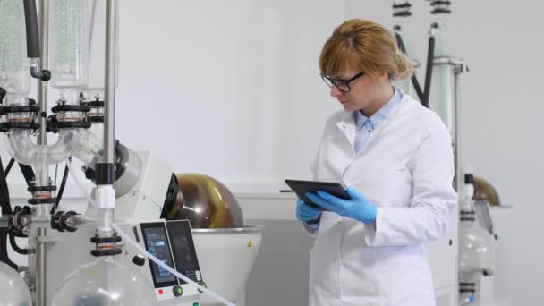Cbd油抽出中に研究室でタブレットを入力する科学者 彼女はゴム手袋を着用し Rotavaporと呼ばれる緑のコンデンサーと回転気化器の横に立っている — ストック動画