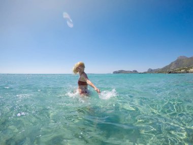 Girit adasındaki sahilde bikinili, sığ turkuaz suya atlayan özgür ve mutlu bir kadın..