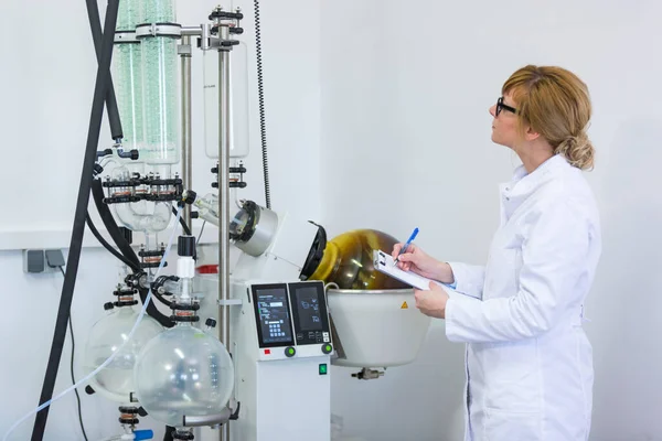 CBD yağ çıkarma sırasında kağıt pano tutan ve rotasyonel buharlaştırıcıyı kontrol eden bir bilim adamı. Makine yeşil yoğunlaştırıcıya ve CBD kenevir yağı çıkarma işleminde olan döner mataraya sahiptir..