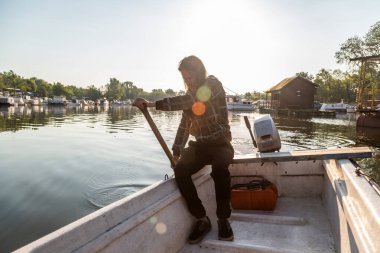 Beyaz orta yaşlı adam eski beyaz teknesinde kürek çekiyor. Gün doğumunda nehir marinasında. Kürek çekmek için bu balıkçı eski tahta kürekleri kullanıyor. Etrafında kayıklar ve yüzen evler ve arka planda ağaçlar var..