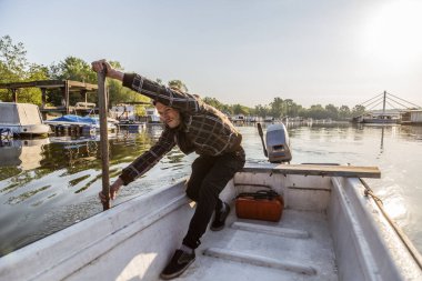 Beyaz orta yaşlı adam eski beyaz teknesinde kürek çekiyor. Gün doğumunda nehir marinasında. Kürek çekmek için bu balıkçı eski tahta kürekleri kullanıyor. Etrafında kayıklar ve yüzen evler ve arka planda ağaçlar var..