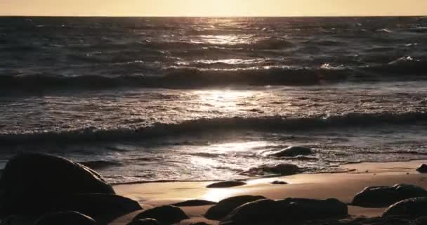 La costa arenosa con el maremoto al atardecer, las piedras, la arena, las olas, nadie, el paisaje, la tranquilidad, el sol cae — Vídeo de stock