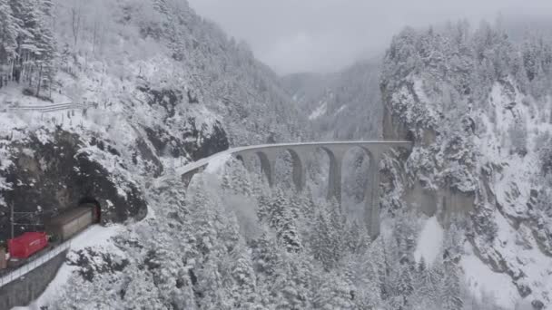 Vista aérea del viaducto Landwasser con tren de carga de color rojo en invierno, El tren se mueve a un túnel, hito de Suiza, nevando, dron vuela hacia adelante — Vídeos de Stock