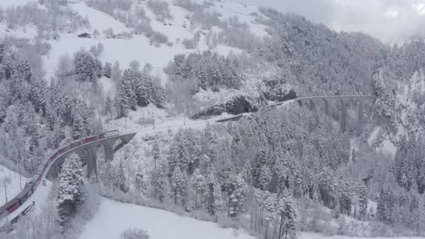 Luftbild des berühmten Zuges von roter Farbe Glacier Express im Winter, Der Zug bewegt sich zum Landwasserviadukt und Tunnel, Wahrzeichen der Schweiz, schneit, Drohne fliegt vorwärts — Stockvideo