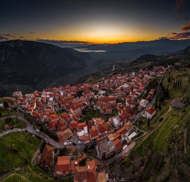 Delphi, Yunanistan, Korint Körfezi 'nin havadan görünüşü, turuncu bulutlar, katmanlı tepeler ve ön planda çatılar.