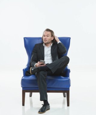 Siyah takım elbiseli yakışıklı adam mavi bir sandalyede oturuyor, üzgün, uzun saçlı, beyaz gömlekli, iş adamı, çok şık, akıllı telefona bakıyor.